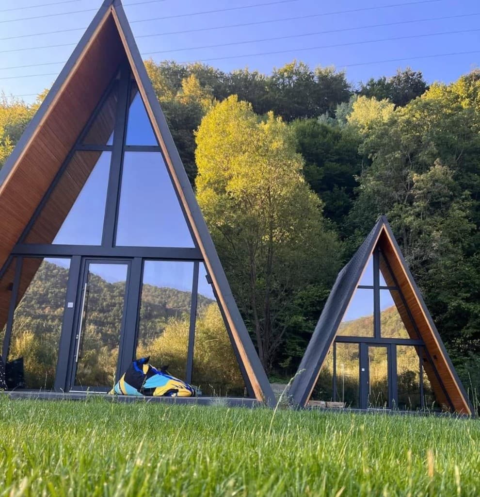 Două cabane A-Frame — structură metalică și fațadă din sticlă, în natură